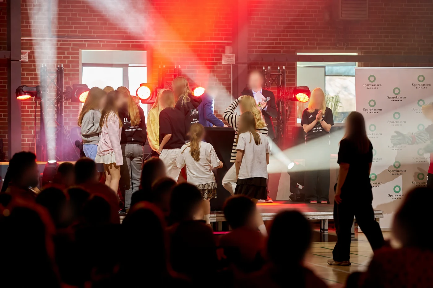 An indoor event with a group on stage, red lighting and white beams.